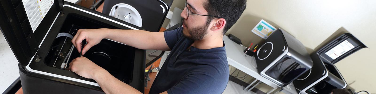 Male student in front of 3D printer, making adjustments in room full of 3D printers.